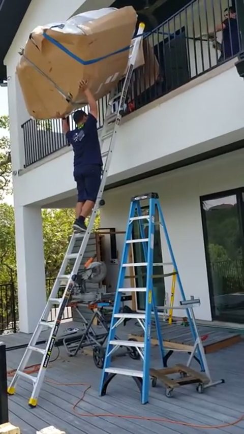 mover on a ladder hoisting a paper-wrapped sectional up to a balcony