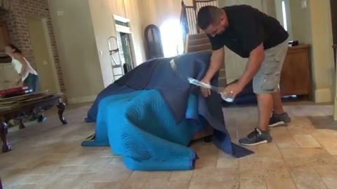 mover shrink-wrapping a blanket-covered sofa in a home, face visible