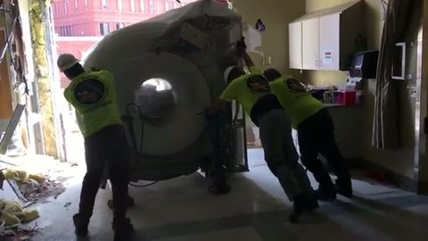 movers pushing a shrink-wrapped MRI scanner through a clinic doorway